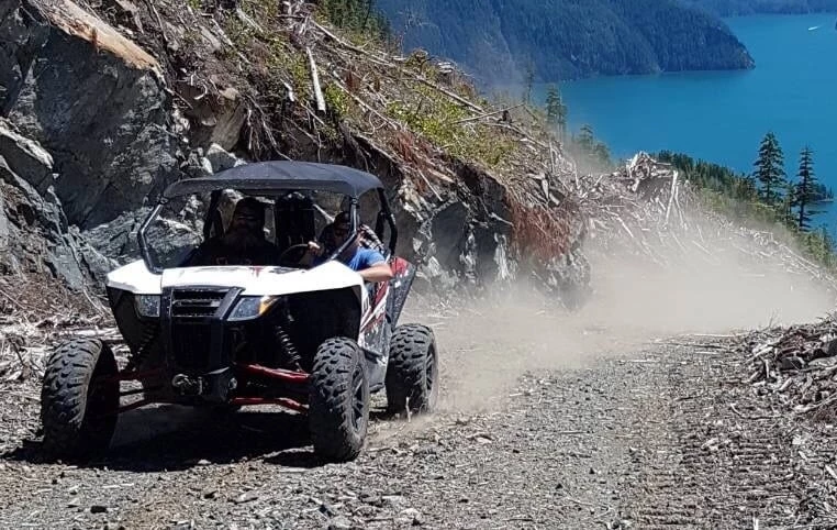 An off-road vehicle with two people is driving on a dusty trail near a forested cliff with a blue water visible in the background.