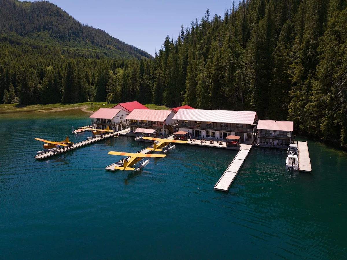 An aerial view of a serene Nootka Sound Resort buildings and multiple seaplanes is surrounded by dense green forests.