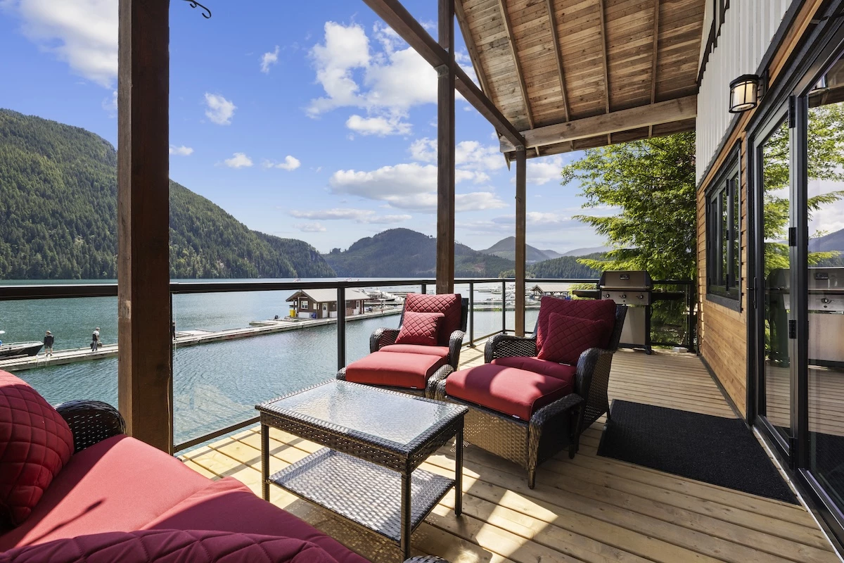 Moutcha Bay Resort Chalet balcony with comfortable red-cushioned chairs, a glass table, barbecue grill, overlooking a dock with boats against a backdrop of mountains.