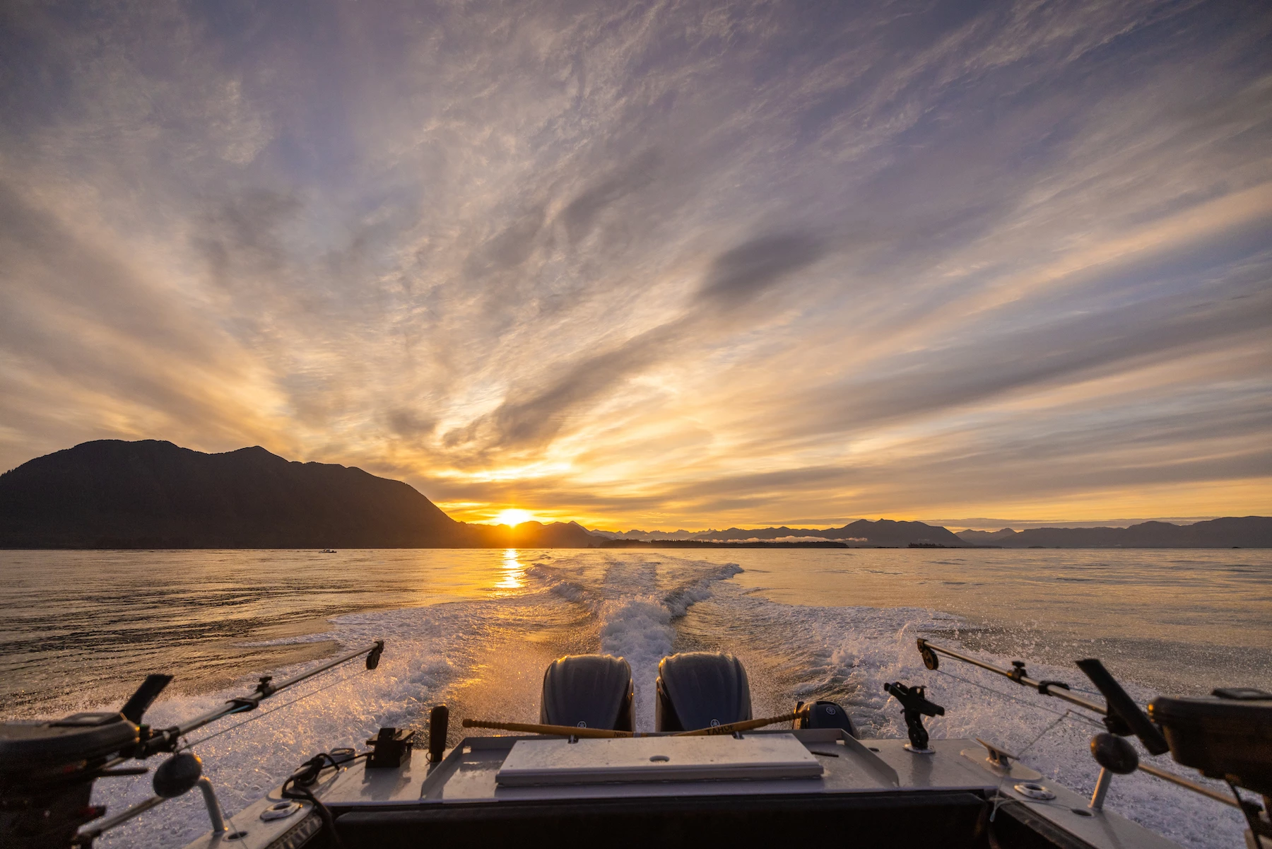 A beautiful sunset viewed from a speeding boat's stern, showing wake trails, fishing rods, mountains in the distance, and a dramatic sky.