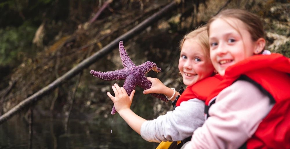 Two children in life jackets are outdoors, excitedly holding a purple starfish near water, with natural scenery in the background.