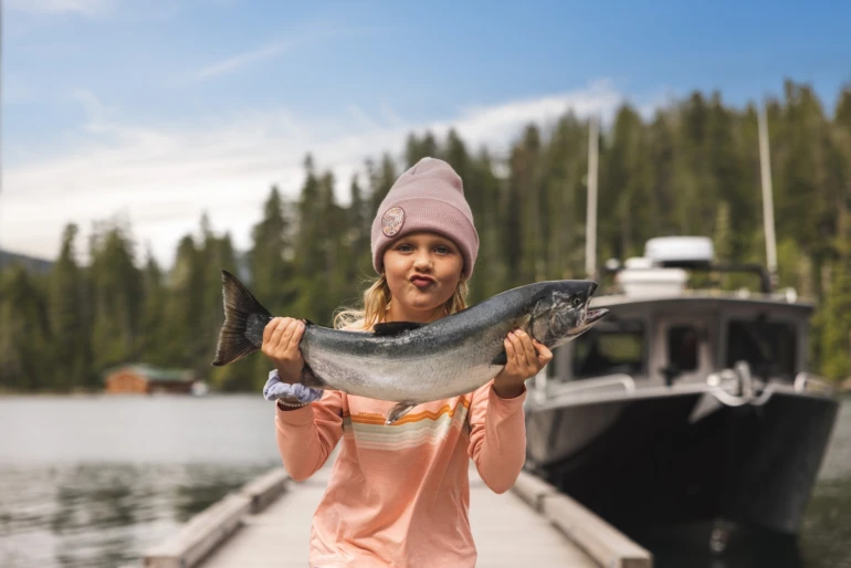 A child stands on a wooden dock, proudly holding a large salmon, with a boat and forest in the background. She appears happy and accomplished.