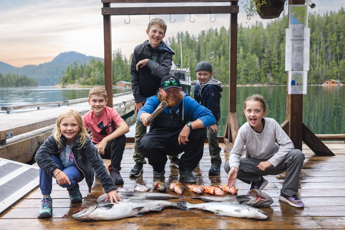 A group of children and a guide with a beard pose happily on a dock with fresh fish, surrounded by a calm water and mountains.