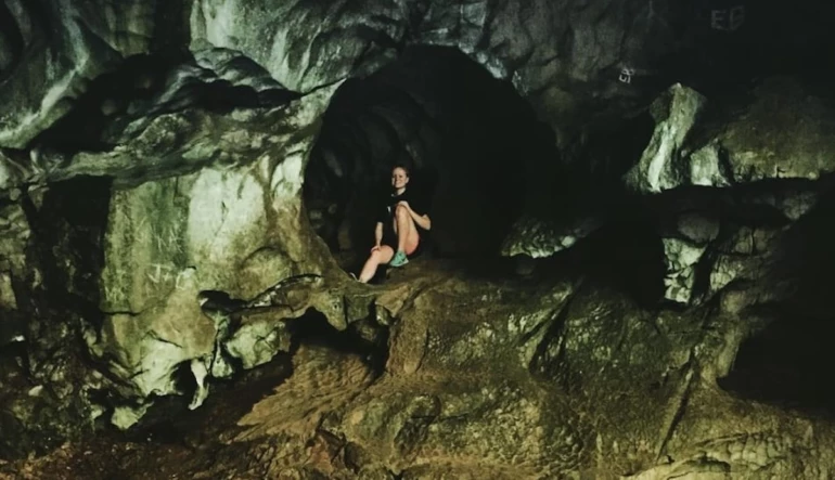 A person sits on a rocky surface inside a dark cave with uneven walls and a visible entrance in the background. The atmosphere is shadowy and mysterious.
