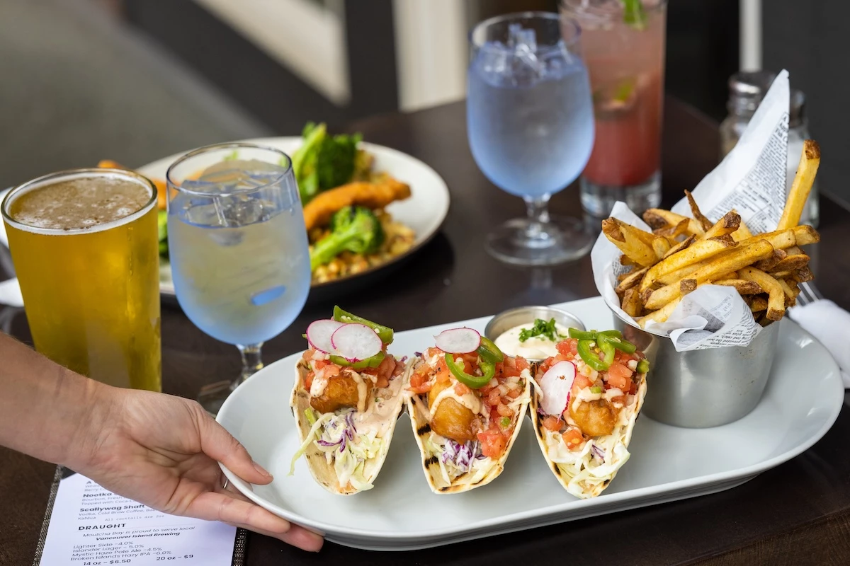 A person is serving a plate of tacos accompanied by a side of crispy fries, cold beverages, a beer, and another dish in the background at Conuma Grill at Moutcha Bay Resort