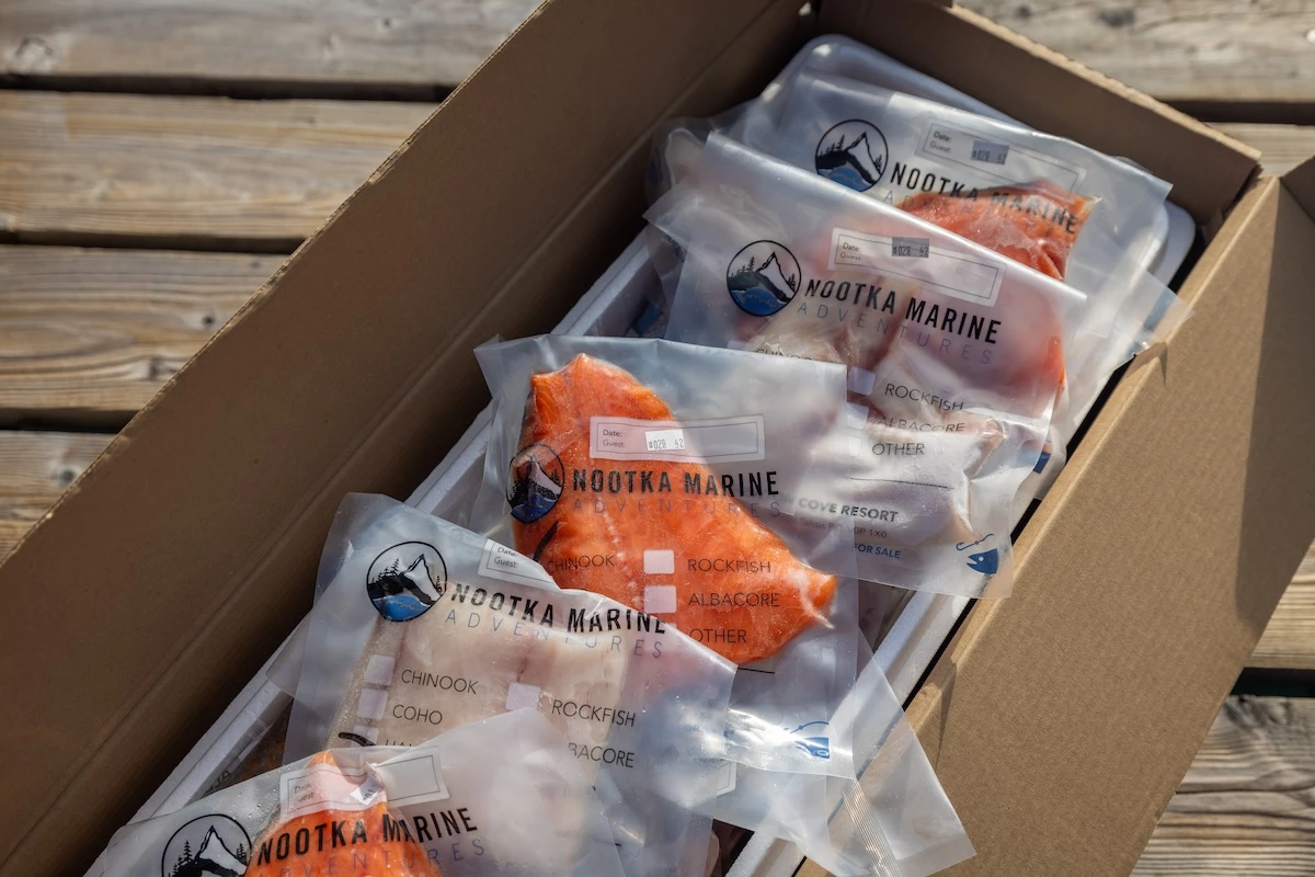 A cardboard box contains multiple vacuum-sealed packages of fish, labeled with types like "Chinook," "Coho," and "Rockfish," branded as "Nootka Marine."