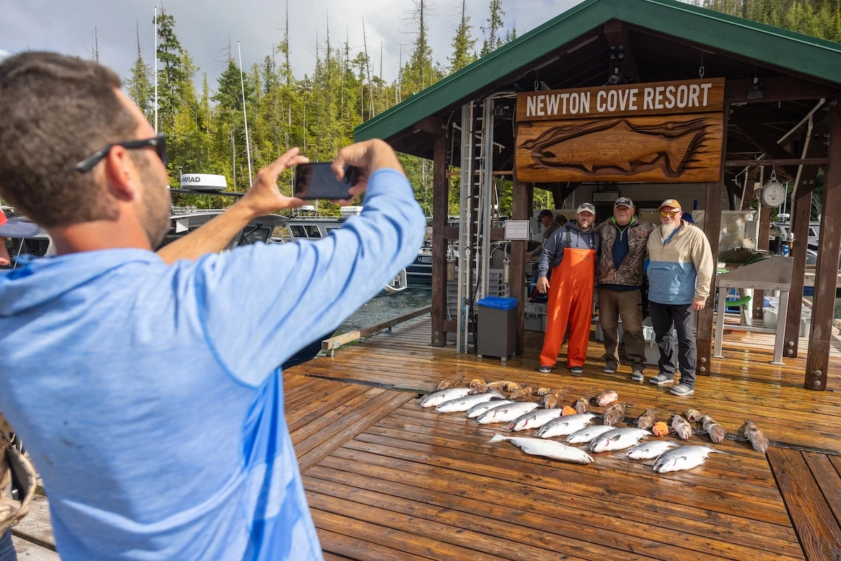 A guest is photographing three guests at a wooden dock by "Newton Cove Resort" sign, standing with a catch of fish displayed on the ground.