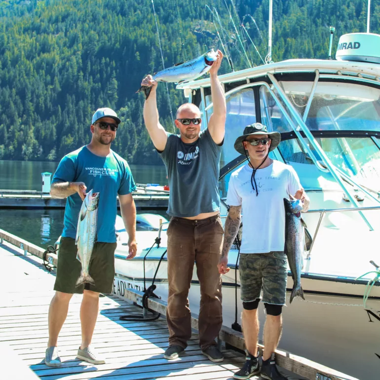Three guest are standing on a dock beside a boat, smiling and holding fish they caught, with a forested hill in the background.