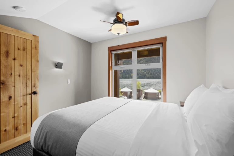 Moutcha Bay Resort lodge bedroom with a large bed, wooden door, ceiling fan, and window overlooking a serene water and mountains in the background.