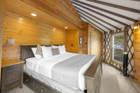 A cozy Moutcha Bay Resort yurt bedroom with wood paneling, a sloped ceiling, a large bed with white bedding, a window with a nature view, and a standing fan.