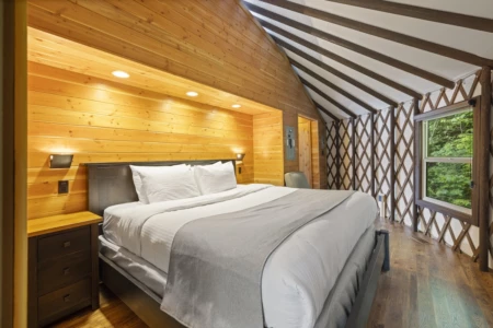 Cozy Moutcha Bay Resort yurt bedroom with a large bed, wooden walls and ceiling, decorative beams, ambient lighting, and a window with a view of green foliage.