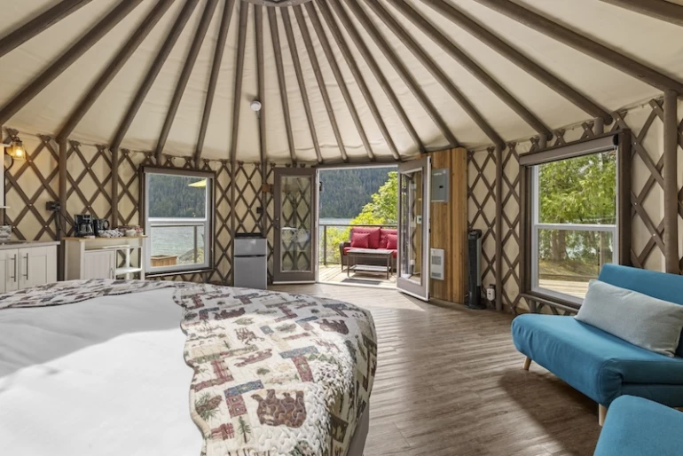 Spacious yurt interior at Moutcha Bay Resort with large bed, patterned quilt, hardwood floors, natural light, and scenic view of a water through glass doors leading to a balcony.