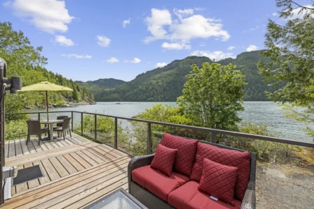 A serene waterside deck with a red sofa, umbrella-shaded chairs, and a stunning view of forested hills under a blue sky with fluffy clouds.