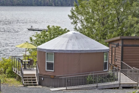 A yurt at Moutcha Bay Resort with a conical roof is connected to a wooden structure overlooking water with a dock, amidst trees, under a cloudy sky.