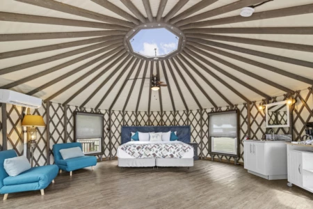 Spacious Moutcha Bay Resort yurt interior at with a radial wooden beam ceiling, a skylight, a large bed, blue chairs, and a kitchenette area.
