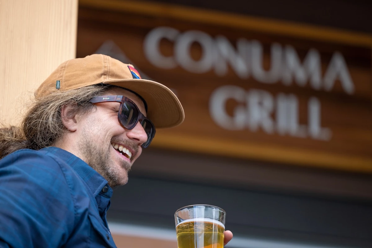 A smiling guest wearing a hat and sunglasses holds a glass of beer, with a blurred sign reading "CONUMA GRILL" in the background.