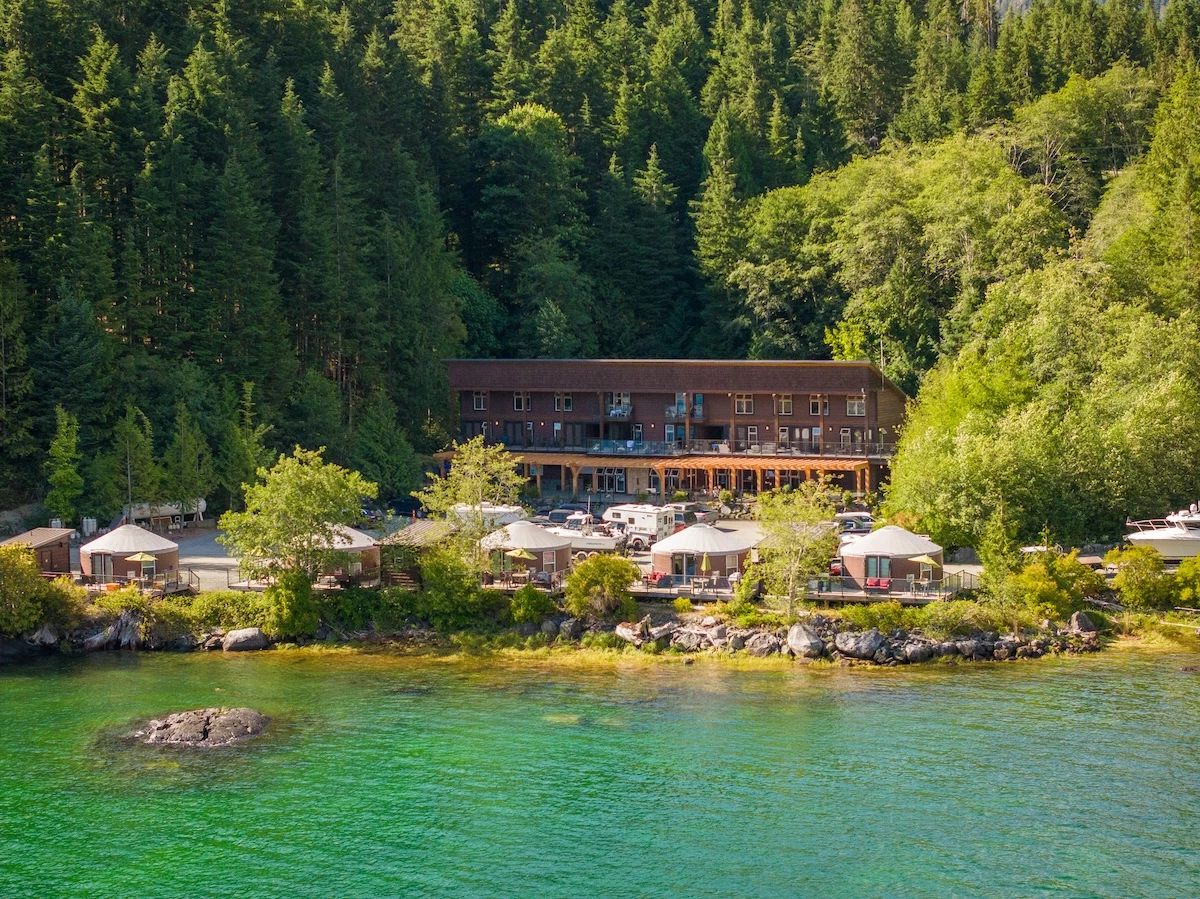 Aerial view of a lakeside resort with yurts, a large lodge, surrounded by dense forests, under a clear sky. Crystal-clear water shimmers on the foreground.