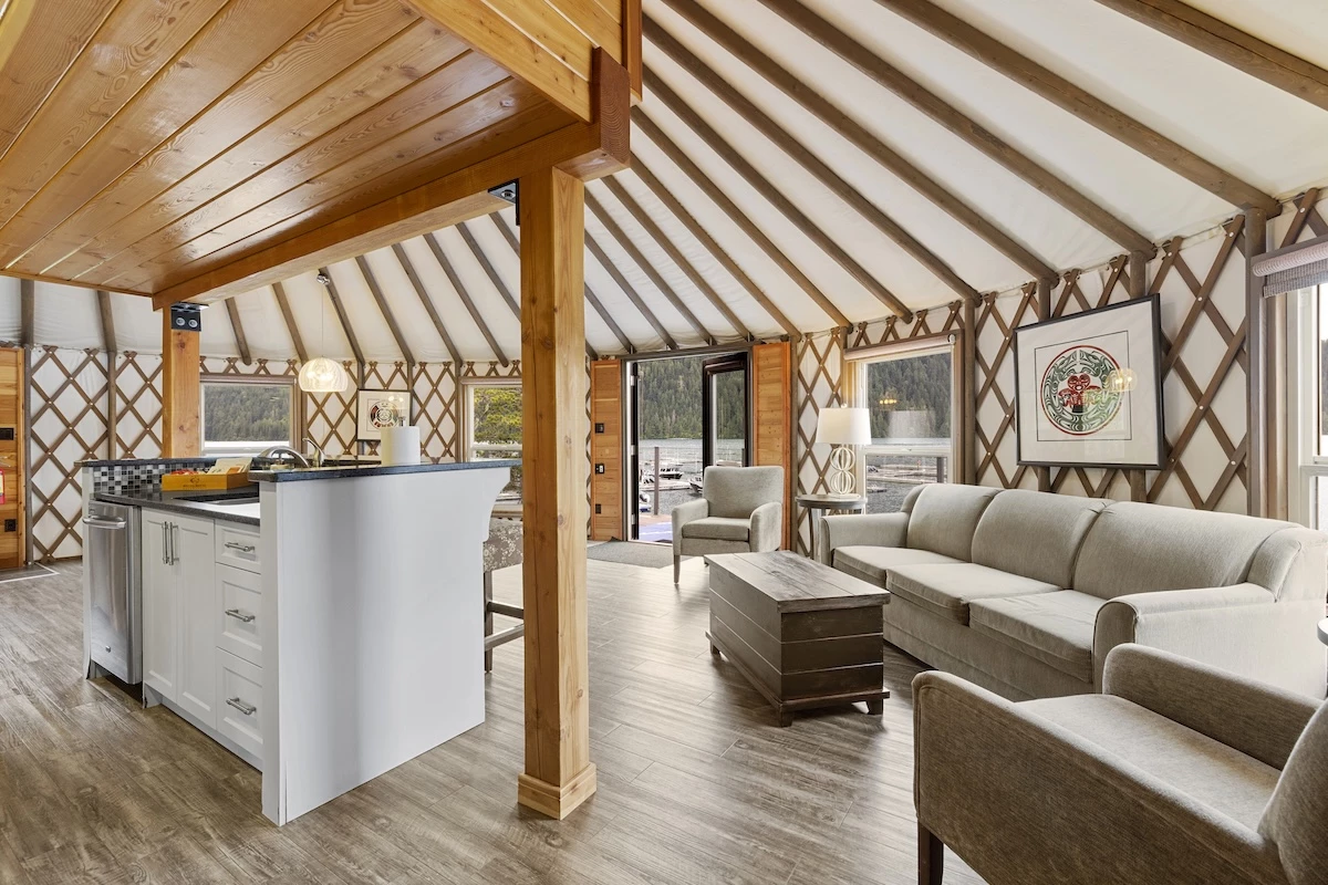 An open interior of a Yurt at Moutcha Bay Resort with wooden beams, a white kitchen island, beige sofas, and a crisscross wall pattern. Large windows provide natural light.
