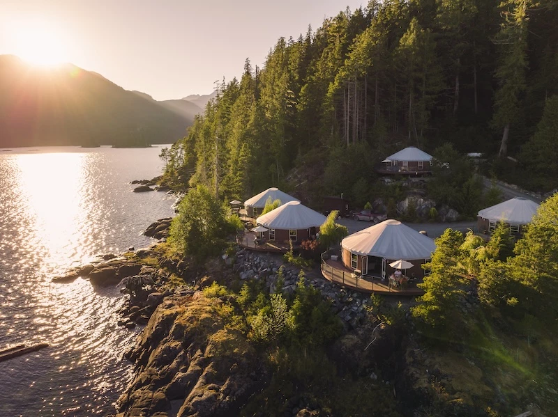 A serene waterfront setting at Moutcha Bay Resort with yurts nestled among trees, as the setting sun casts a warm glow over the tranquil landscape and sparkling water.