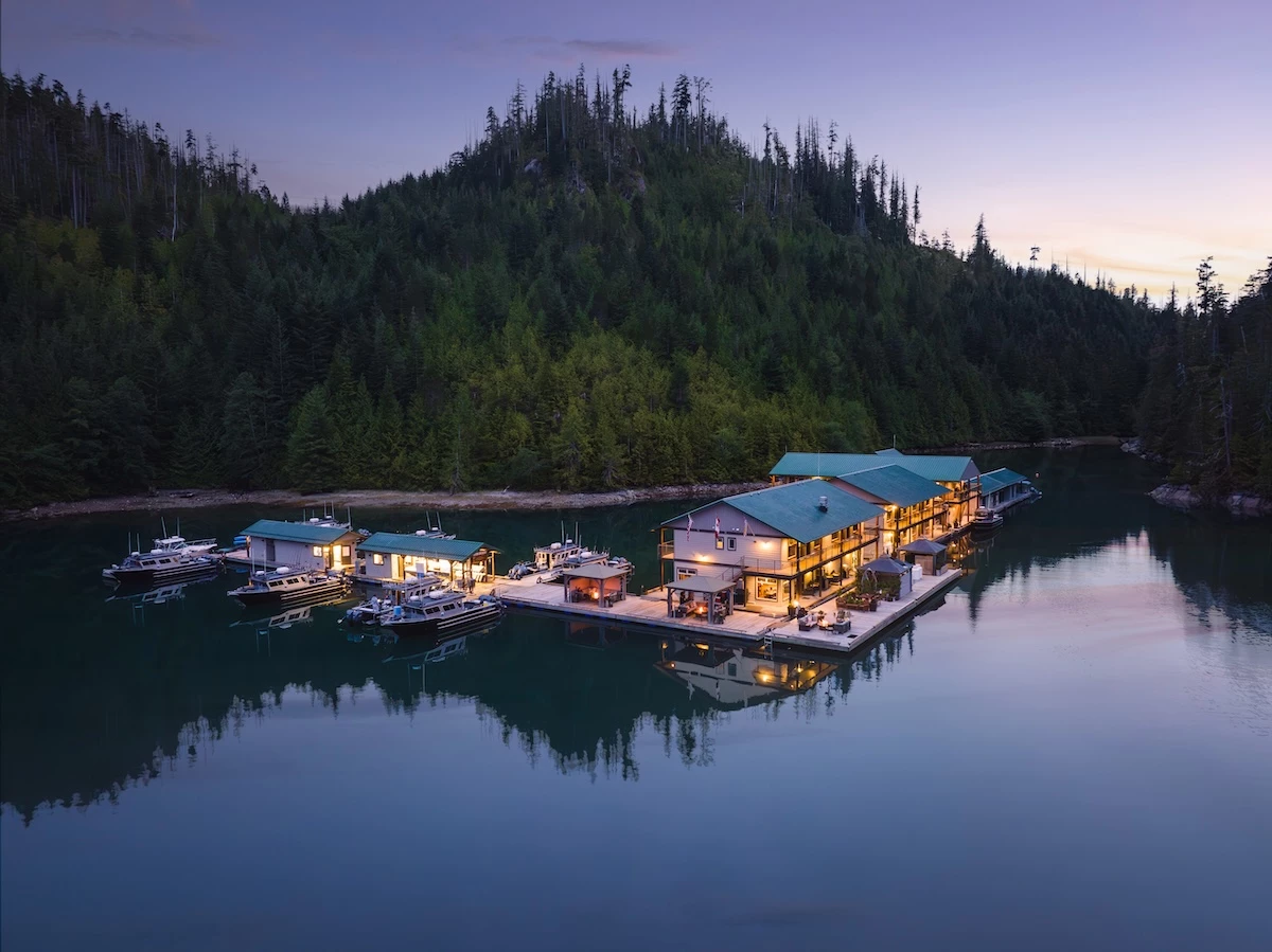 A serene evening at Newton Cove Resort with a lakeside building complex illuminated by warm lights, surrounded by dense forests and moored with several boats.