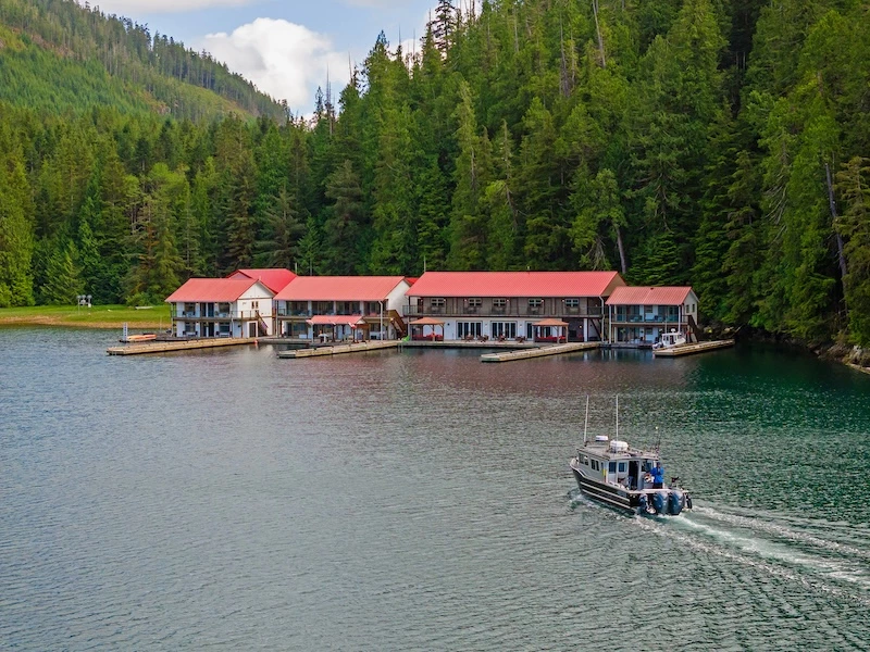 Aerial view of Nootka Sound Resort building with red roofs at the edge of a forested area, with a boat nearby on a serene water.