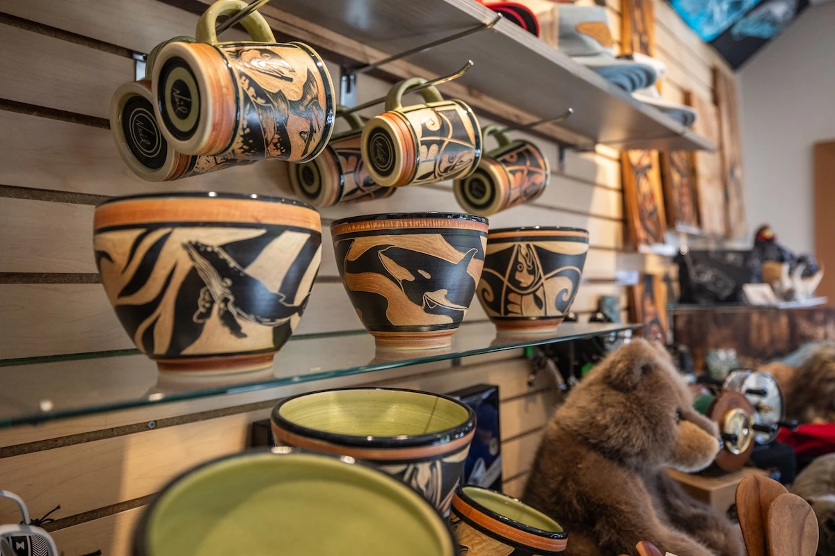 Decorative ceramic pottery with intricate designs is displayed on shelves. Mugs hang above while bowls rest below. A cozy shop atmosphere is depicted.