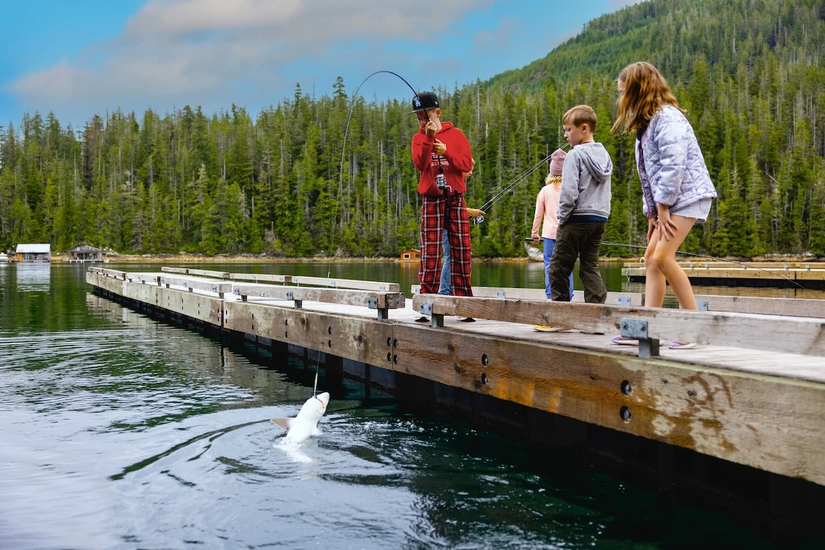 Three kids are fishing from a long wooden dock at Nootka Sound Resort surrounded by a forest.