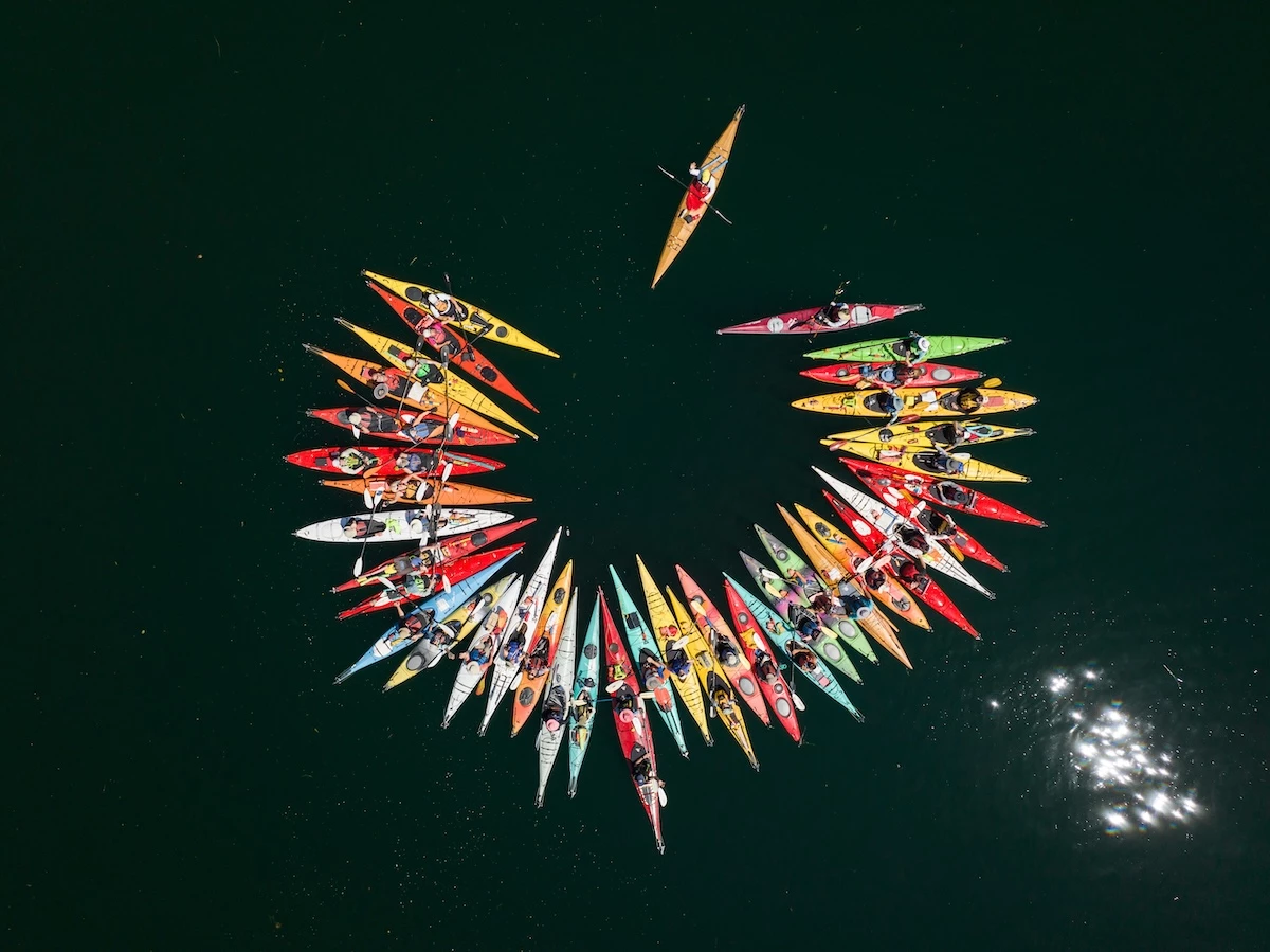 An aerial view of guest in colorful kayaks forming a circle on calm water, with sun reflection to the side.