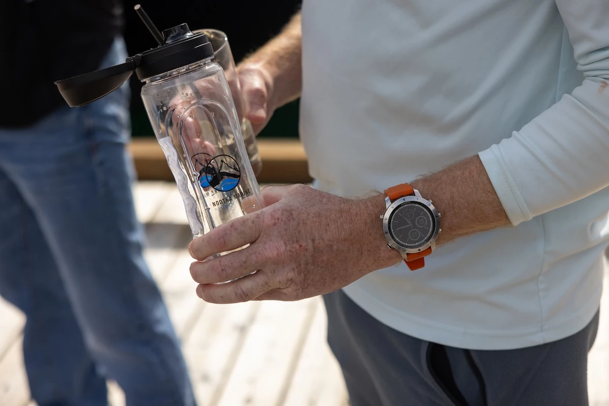 A guest is holding a transparent water bottle with a Nootka Marine Adventures logo, wearing a white long-sleeved shirt and a watch with an orange strap.