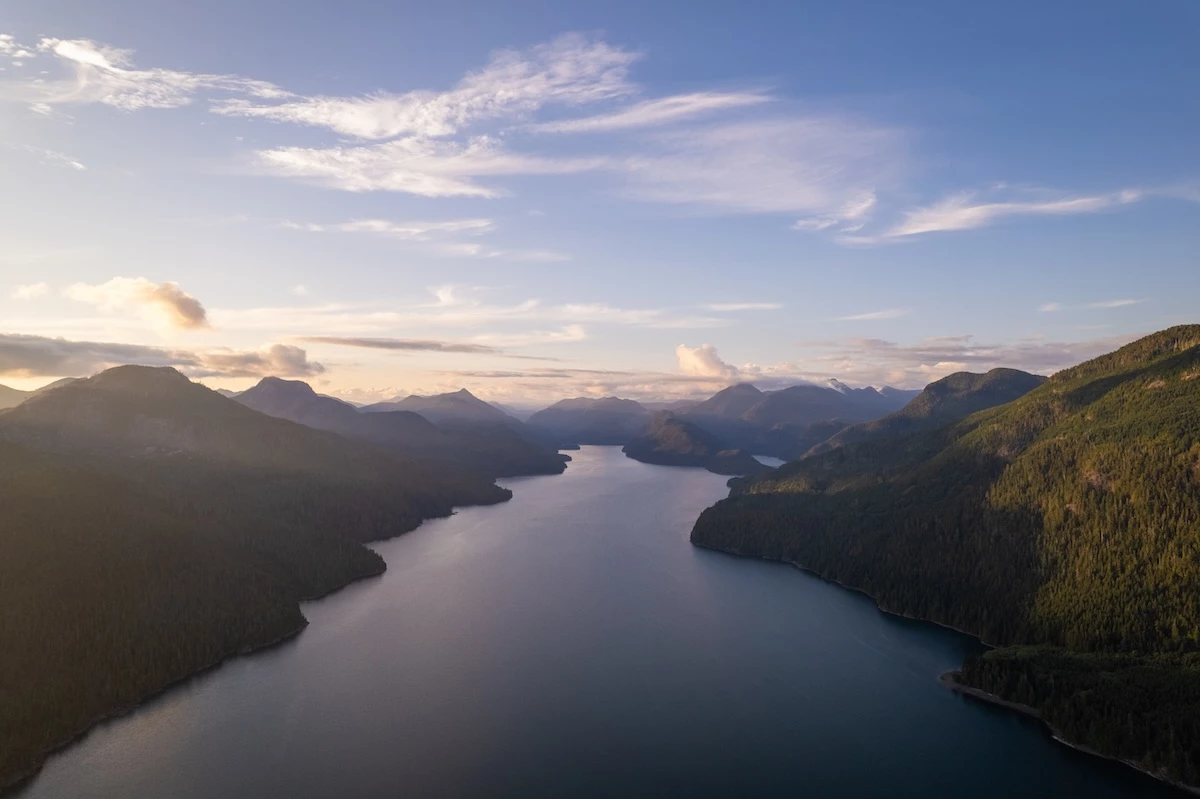 An aerial view of a Nootka Sound nestled among forest-covered hills under a pastel sky with light cloud cover at sunrise or sunset.